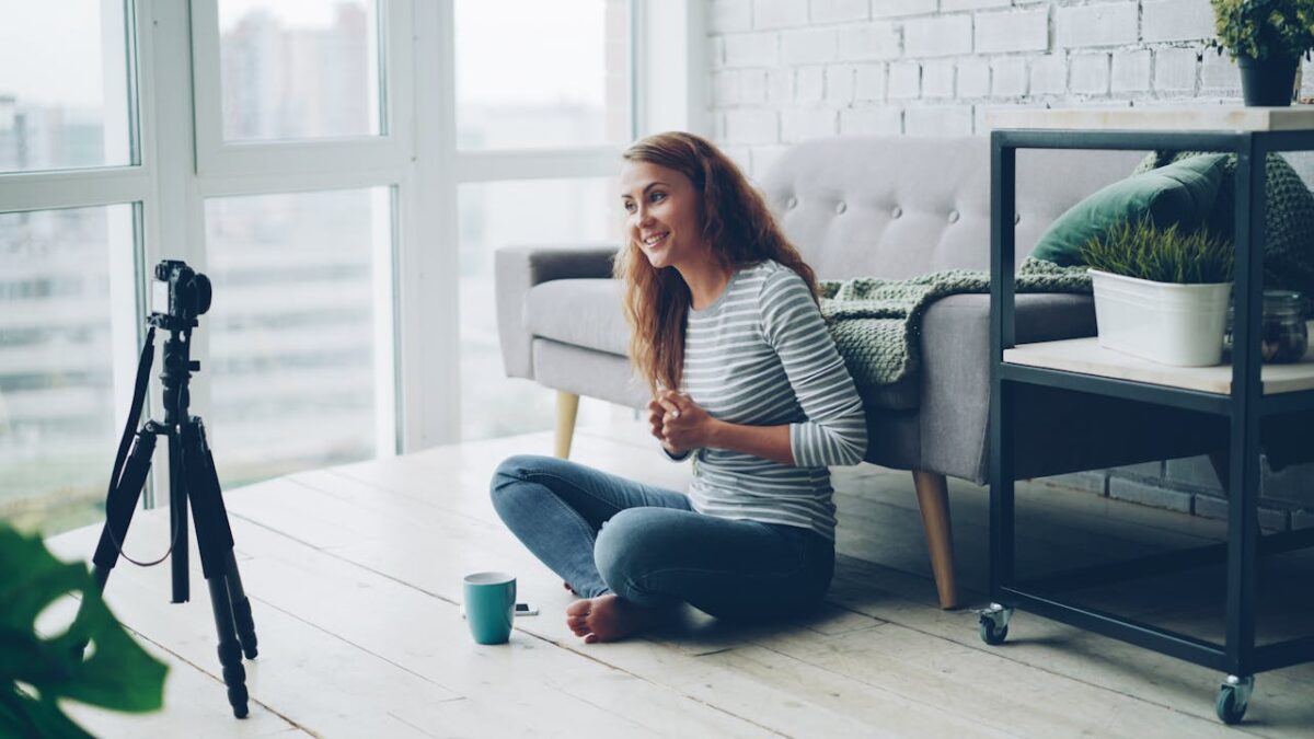 Smiling woman sitting on the floor vlogging with camera tripod in a modern living room.